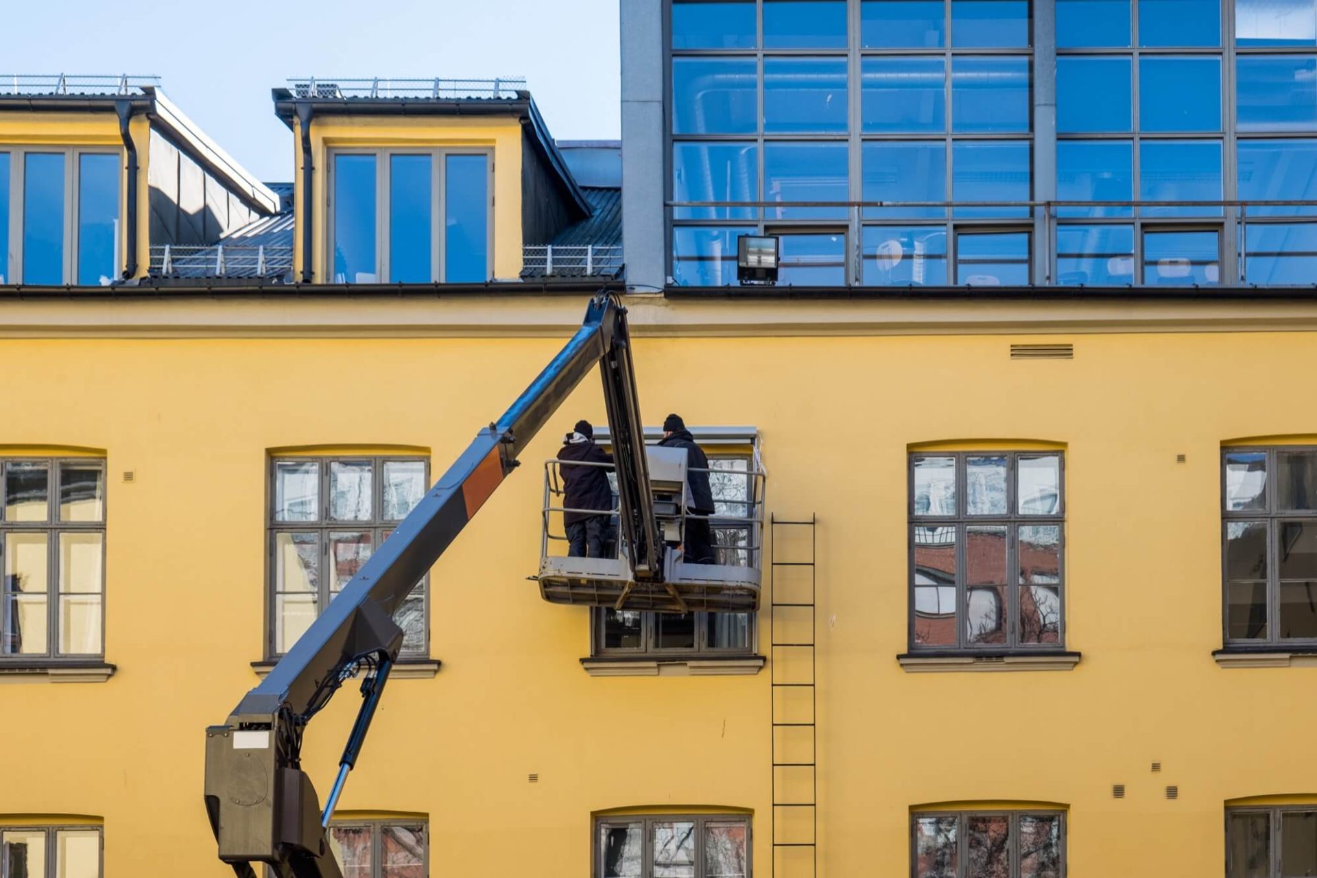 På en solgul byggnad som är under uppfräschning ser vi två arbetare som utför yttre arbeten med hjälp av en skylift