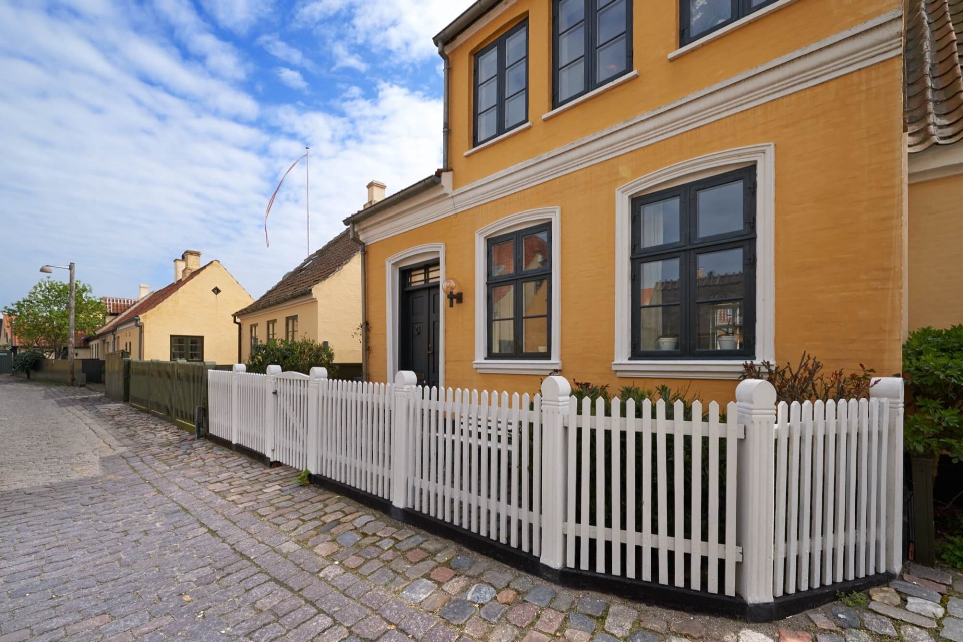 En villa med gul puts, svarta fönster och en vit träport står längs en gata med kullersten